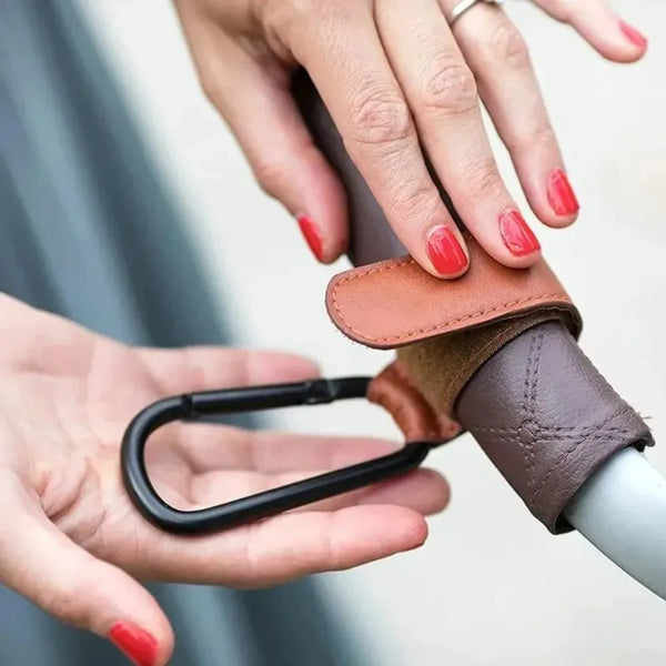 A hand is holding a brown leather stroller hook carabiner accessory.
