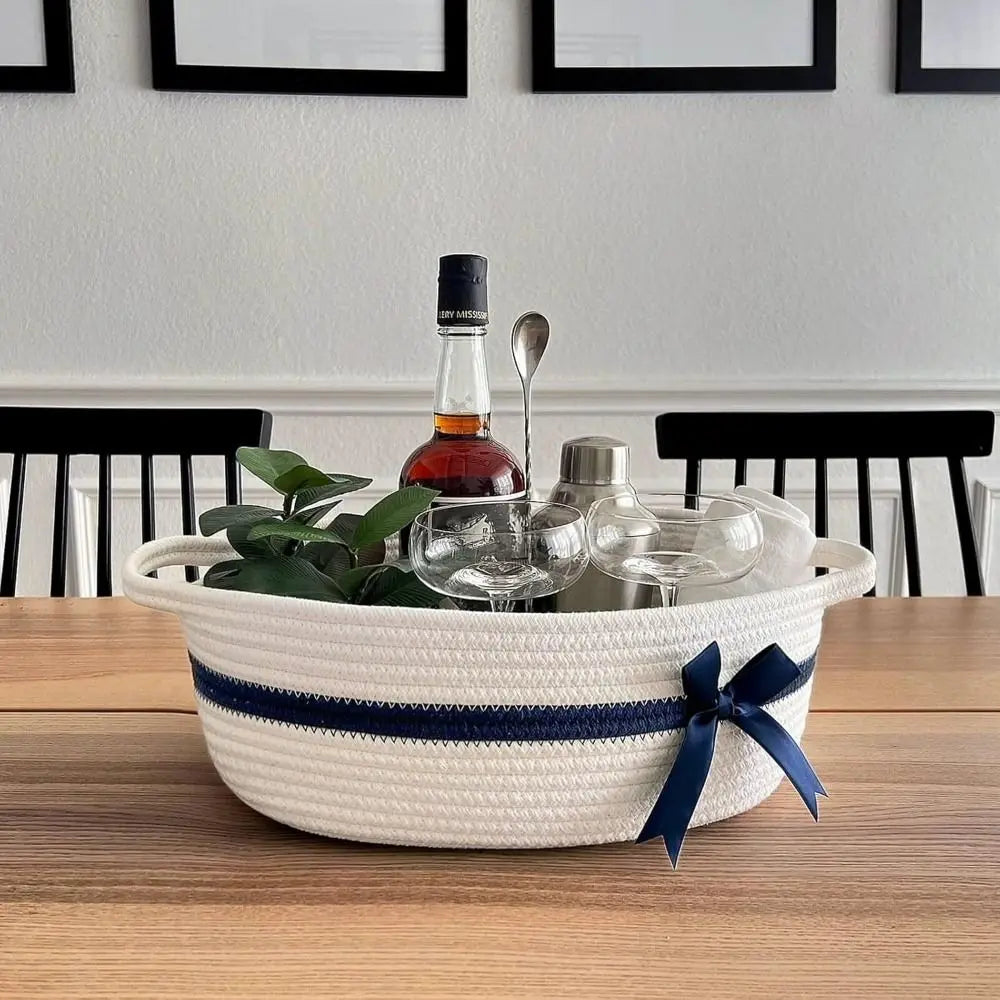 A woven cotton rope storage basket holding a bottle, spoon, and various glassware on a wooden table.