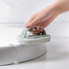 A hand is using a light green magic sponge scrubber with an ergonomic handle to clean the rim of a white toilet bowl.