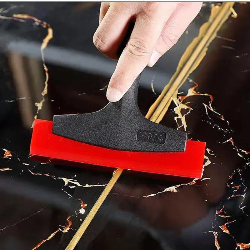 A hand is using a black silicone scraper to fill gaps in grout or cracks on a dark marble surface.