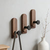 A set of three dark brown walnut wood wall hooks with self-adhesive mounting hardware mounted on a light gray wall.
