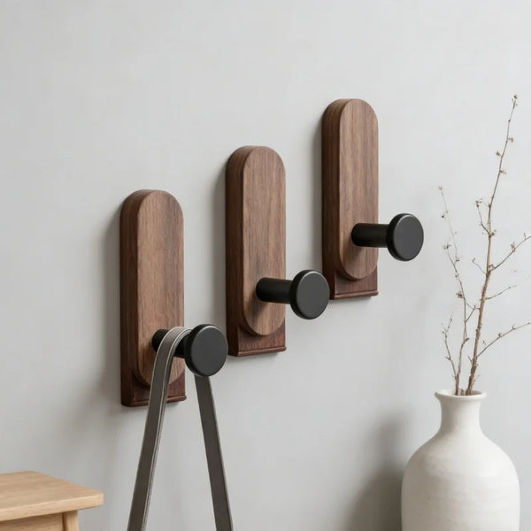 A set of three dark brown walnut wood wall hooks with self-adhesive mounting hardware mounted on a light gray wall.