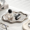 A silver jewelry organizer tray holds a small glass bottle and a pair of eyeglasses on a white surface.