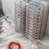 A clear plastic jewelry organizer box filled with multiple small jewelry pieces and a loose red beaded bracelet on a white fl