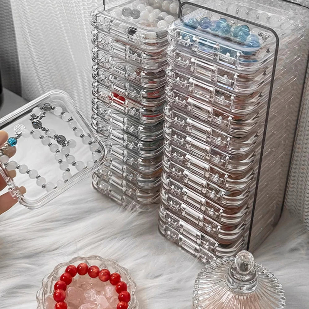 A clear plastic jewelry organizer box filled with multiple small jewelry pieces and a loose red beaded bracelet on a white fl