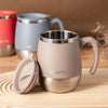 A beige insulated stainless steel travel mug with a silver lid is shown next to other colored mugs on a wooden surface.