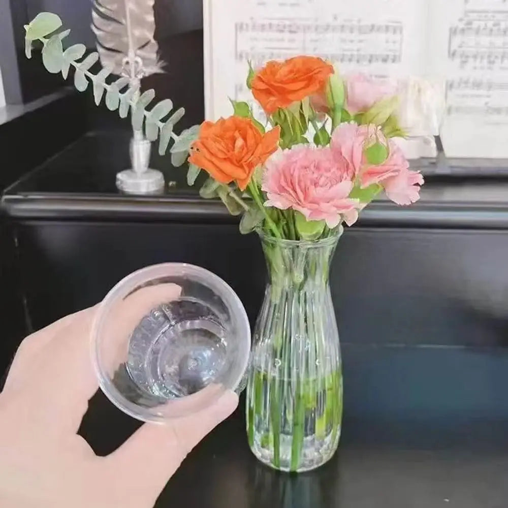 Una mano sostiene un vaso de plástico transparente mientras un jarrón de rosas naranjas y rosas se encuentra sobre una superf