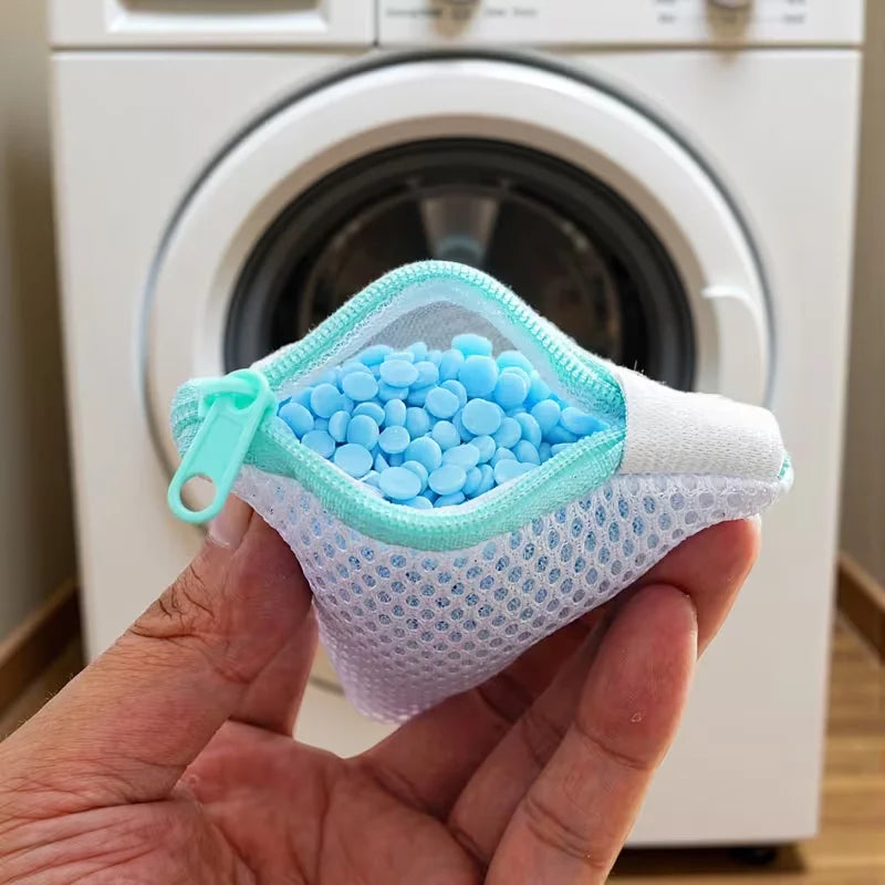 A hand holds a reusable mesh laundry bag filled with blue plastic balls, next to a white washing machine.