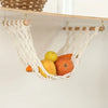 A white macrame fruit hammock made of natural fiber is hanging on a wooden shelf.
