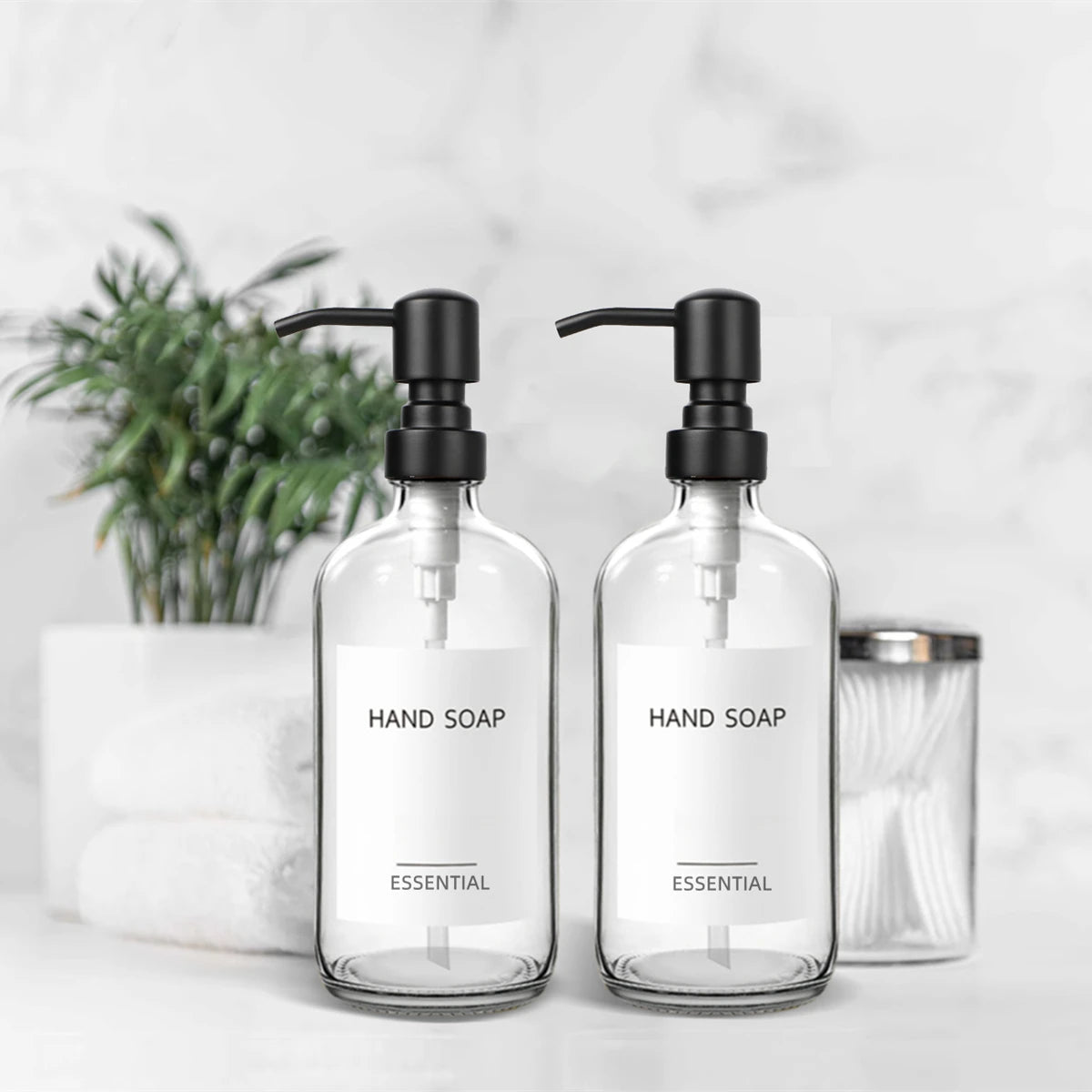 A set of clear glass hand soap dispensers with black pumps and stainless steel accents on a white surface.