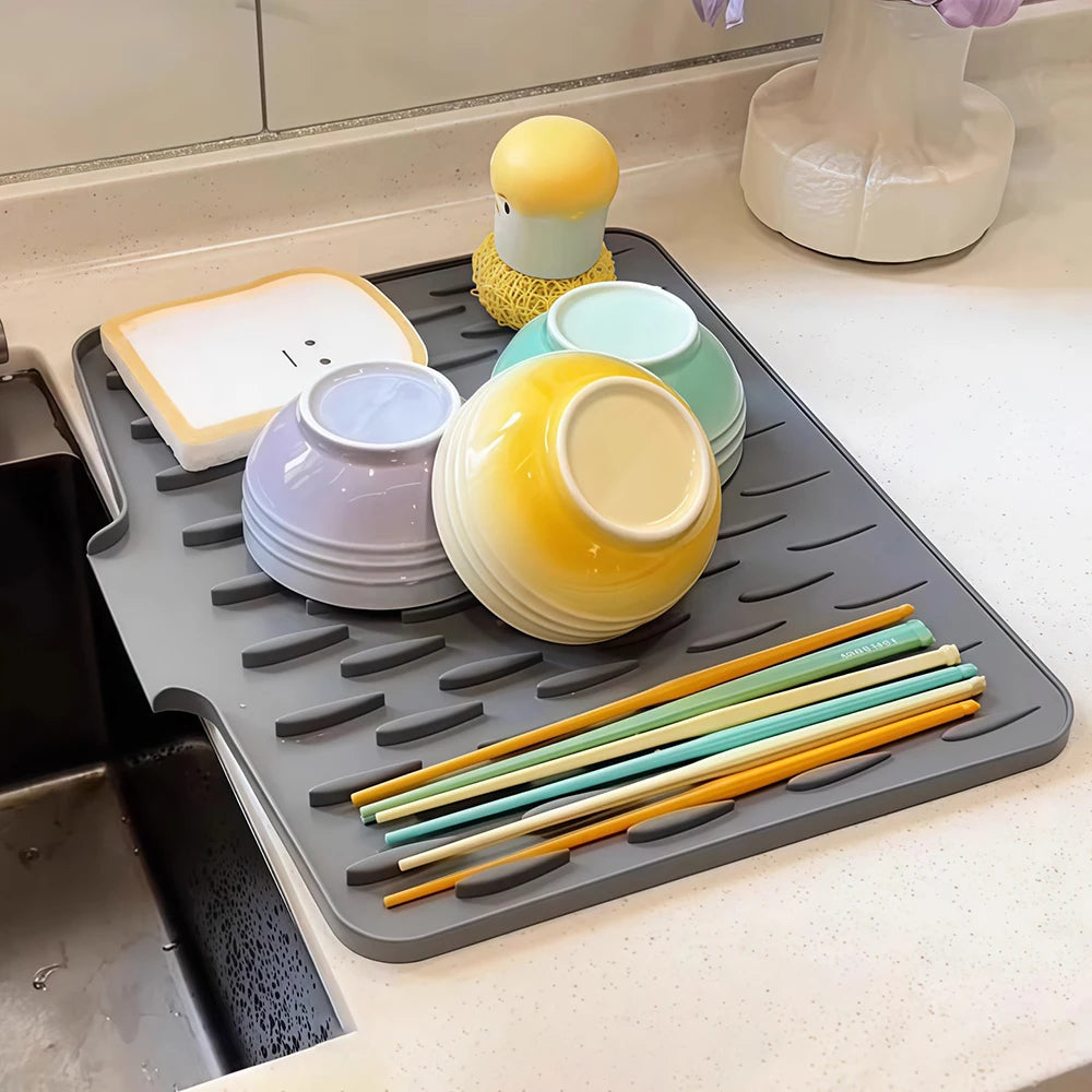 A silicone drain mat with various colored silicone bowls and yellow and green silicone sticks on a countertop.