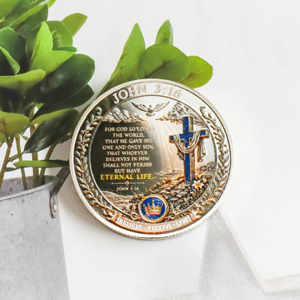 A commemorative coin featuring the Bible verse John 3:16 with a cross and laurel wreath, resting on green foliage.