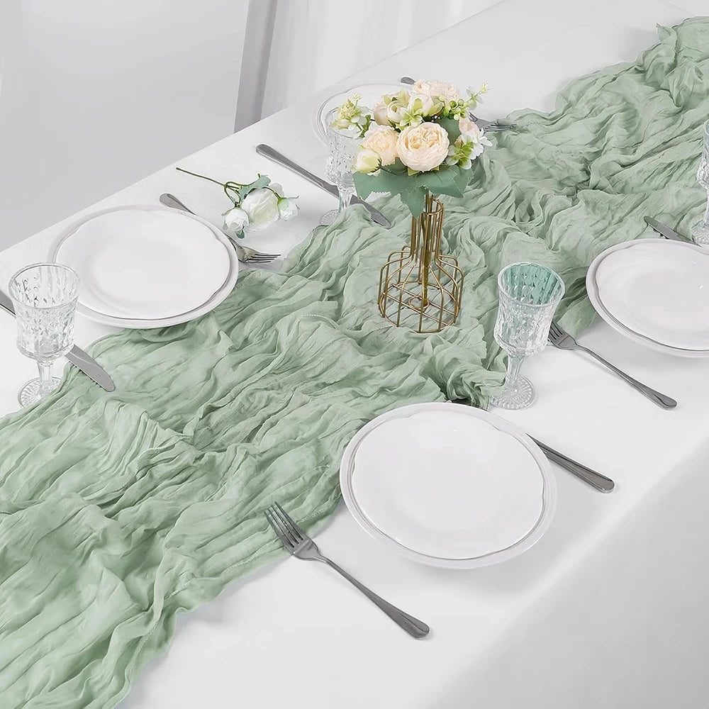A white table setting featuring a light green sheer fabric runner, white plates, silverware, and floral centerpiece.