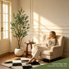 An artificial indoor olive tree in a pot is displayed next to a woman relaxing in an armchair.