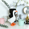 A white goose sculpture with an orange beak and legs is displayed on a white surface next to a potted plant and a teacup.