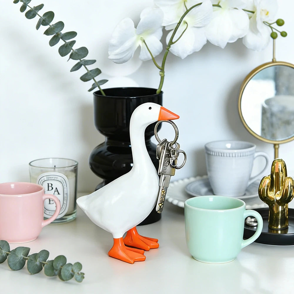 A white goose sculpture with an orange beak and legs is displayed on a white surface next to a potted plant and a teacup.