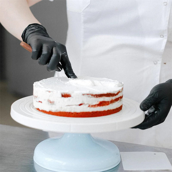 A hand wearing a black nitrile glove is applying frosting to a red cake on a white cake stand.
