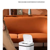 A white touchless sensor trash can is shown on a carpeted floor next to a person's leg.