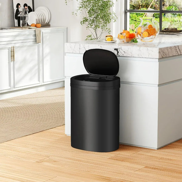 A black automatic touchless trash can sitting on a light wood floor next to a kitchen counter.