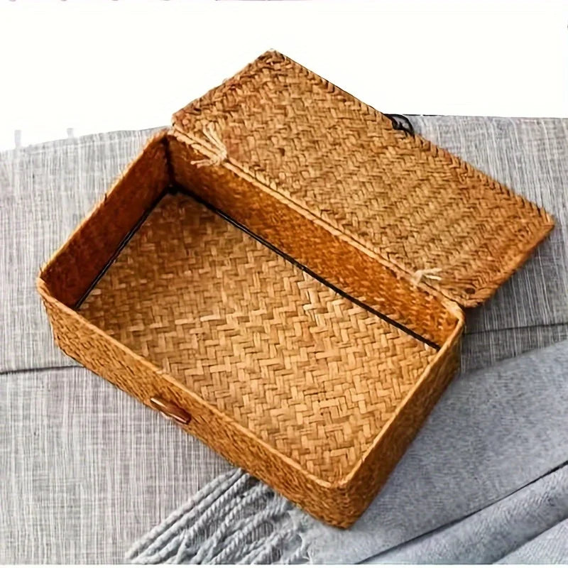 A square handwoven rattan storage basket with a lid resting on a gray fabric surface.