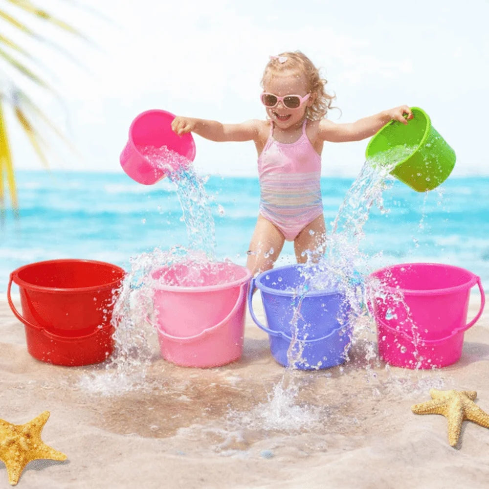A young girl is splashing water from various colored plastic buckets onto the sand near the ocean.