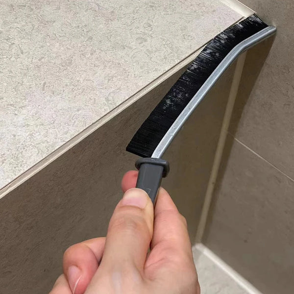 A hand is holding a gray and black grout gap cleaning brush against a tile surface.