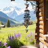 A handcrafted metal wind chime decoration hangs by a window, set against a scenic mountain landscape.