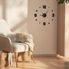 A round black wall clock with Roman numerals hangs on a beige wall next to a light-colored upholstered chair.