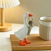 A white goose sculpture with an orange beak and feet holding a silver key chain on a wooden surface.