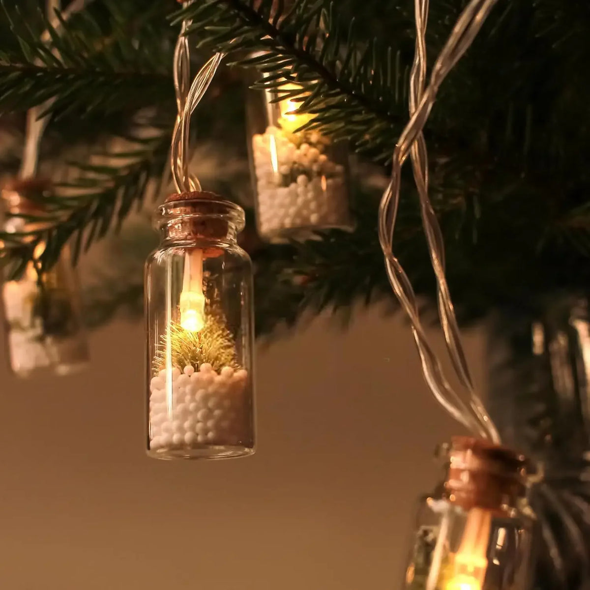 A close-up of a glass bottle candle with a glowing wick and white textured wax, hanging from a green pine branch.