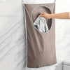 A hand is folding a light gray towel inside a beige fabric laundry basket organizer hanging on a white tiled wall.