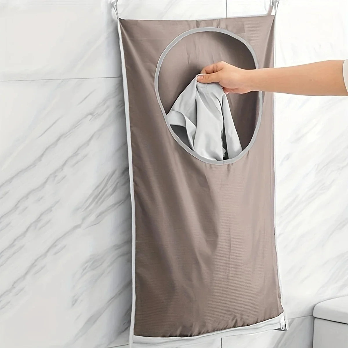 A hand is folding a light gray towel inside a beige fabric laundry basket organizer hanging on a white tiled wall.