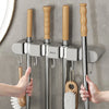 Hands are installing a wall mounted broom holder with wooden handles and metal rods onto a gray tiled wall.