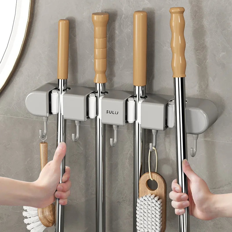 Hands are installing a wall mounted broom holder with wooden handles and metal rods onto a gray tiled wall.