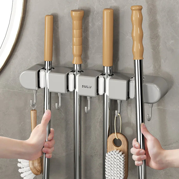 Hands are installing a wall mounted broom holder with wooden handles and metal rods onto a gray tiled wall.