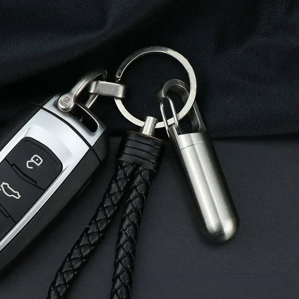 A stainless steel travel pill box and first aid container with a leather cord and keyring.