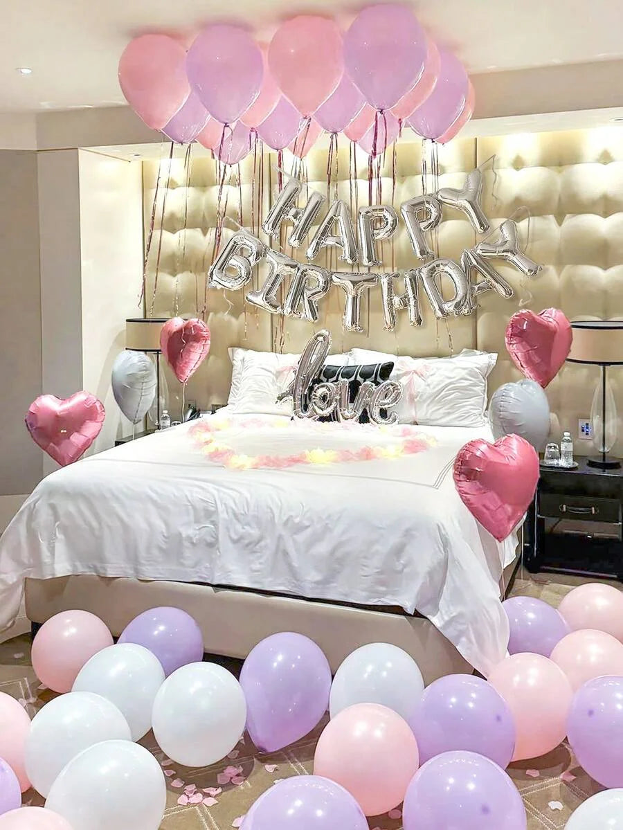 A celebration setup featuring pink and purple foil balloons spelling out Happy Birthday above a bed.