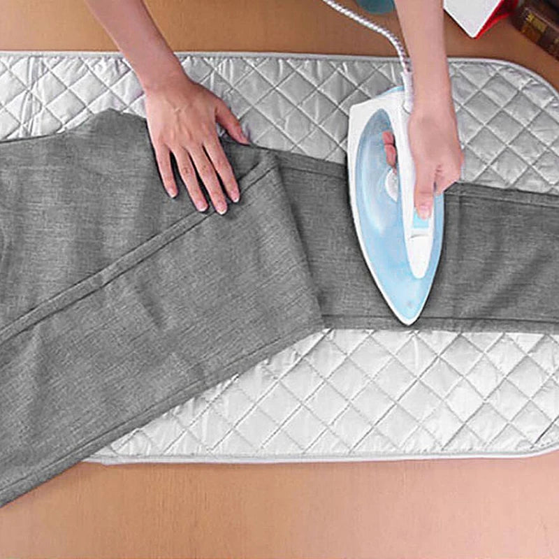 Hands are using a handheld steamer on a gray ironing mat placed on a wooden surface.