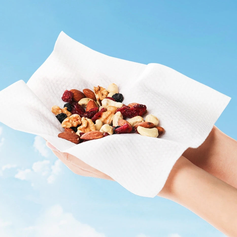 A hand holds a white disposable facial cleansing towel filled with mixed nuts and dried fruit against a blue sky background.