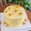 A yellow birthday cake decorated with small yellow bees and white flowers, topped with a gold acrylic topper.