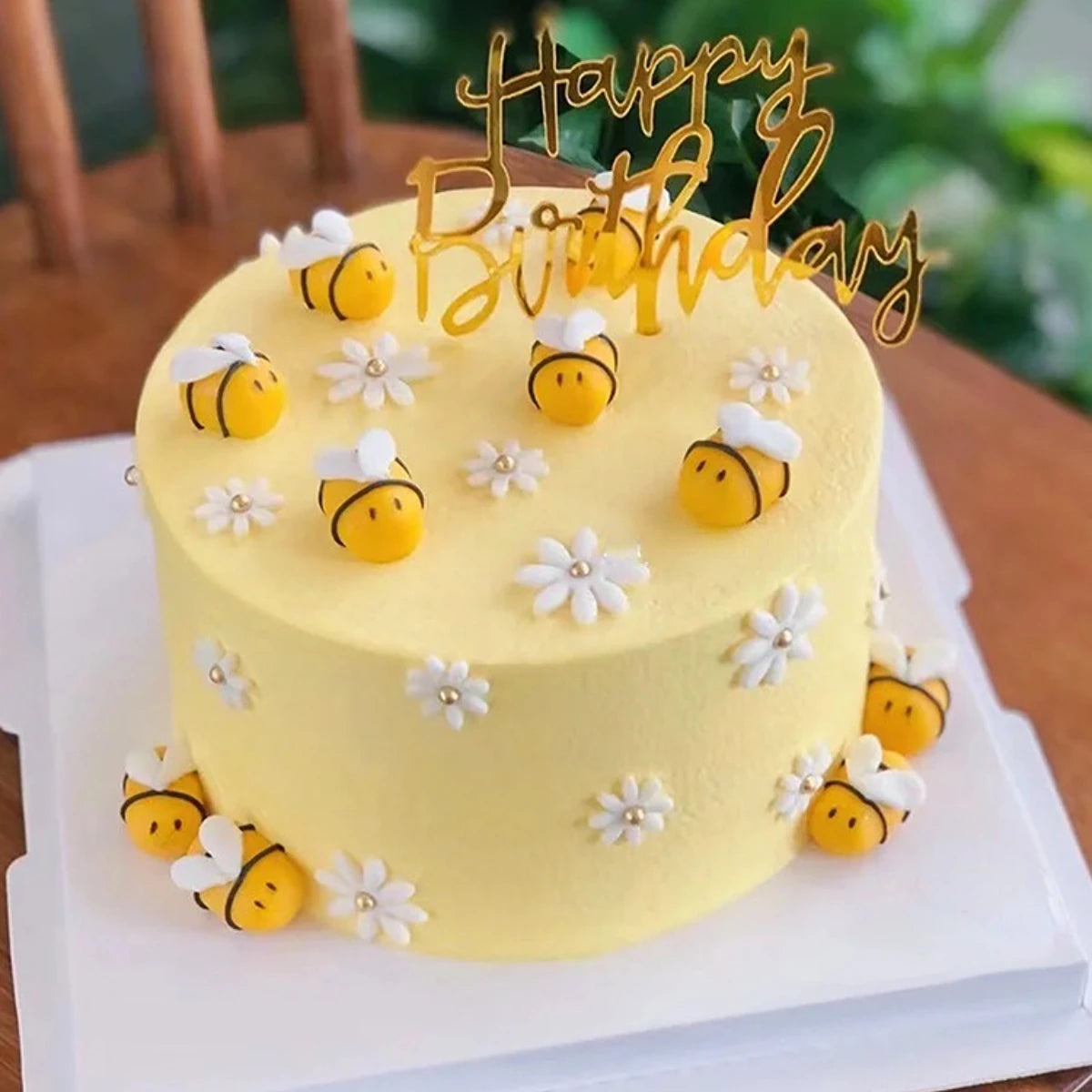 A yellow birthday cake decorated with small yellow bees and white flowers, topped with a gold acrylic topper.