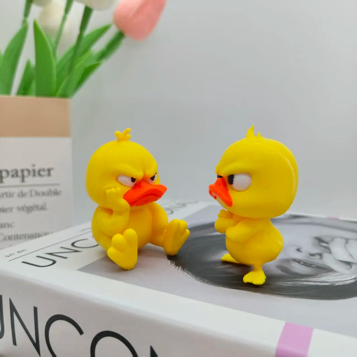 Statue de canard imprimée en 3D jaune décorant une boîte blanche à côté d'un tulipe rose.