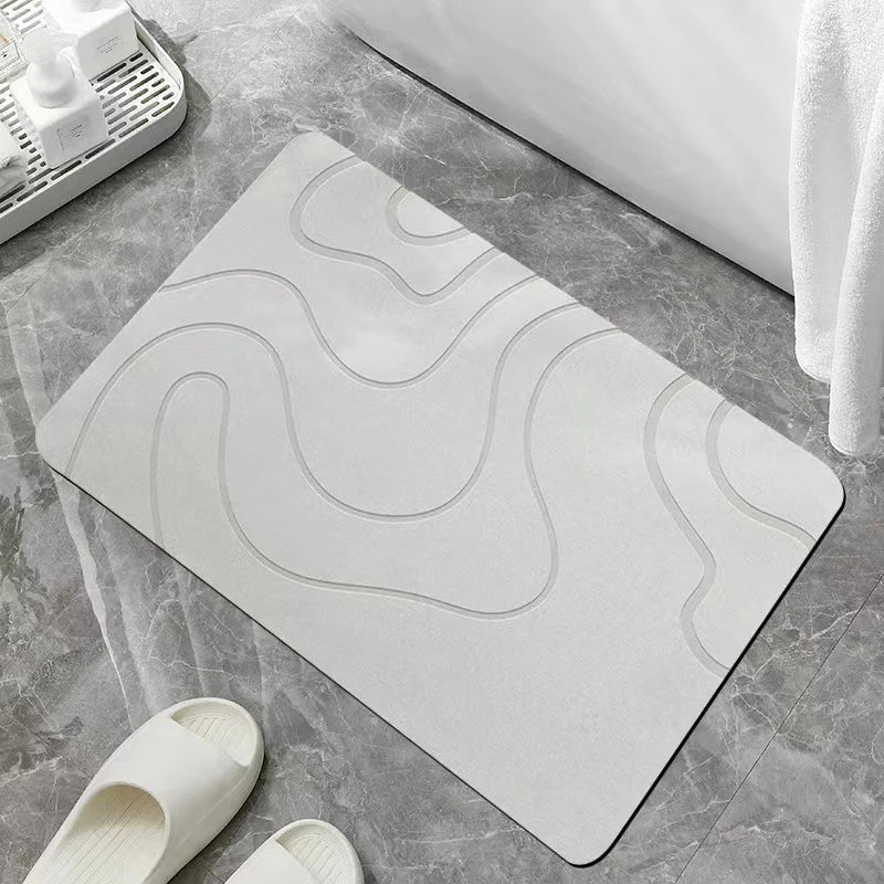 A white bathroom mat with wavy lines is placed on a gray tile floor next to a pair of white slippers.