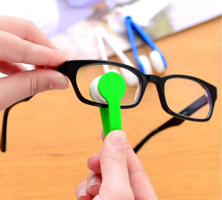 A hand is using a green cleaning tool to wipe the lens of black eyeglasses.