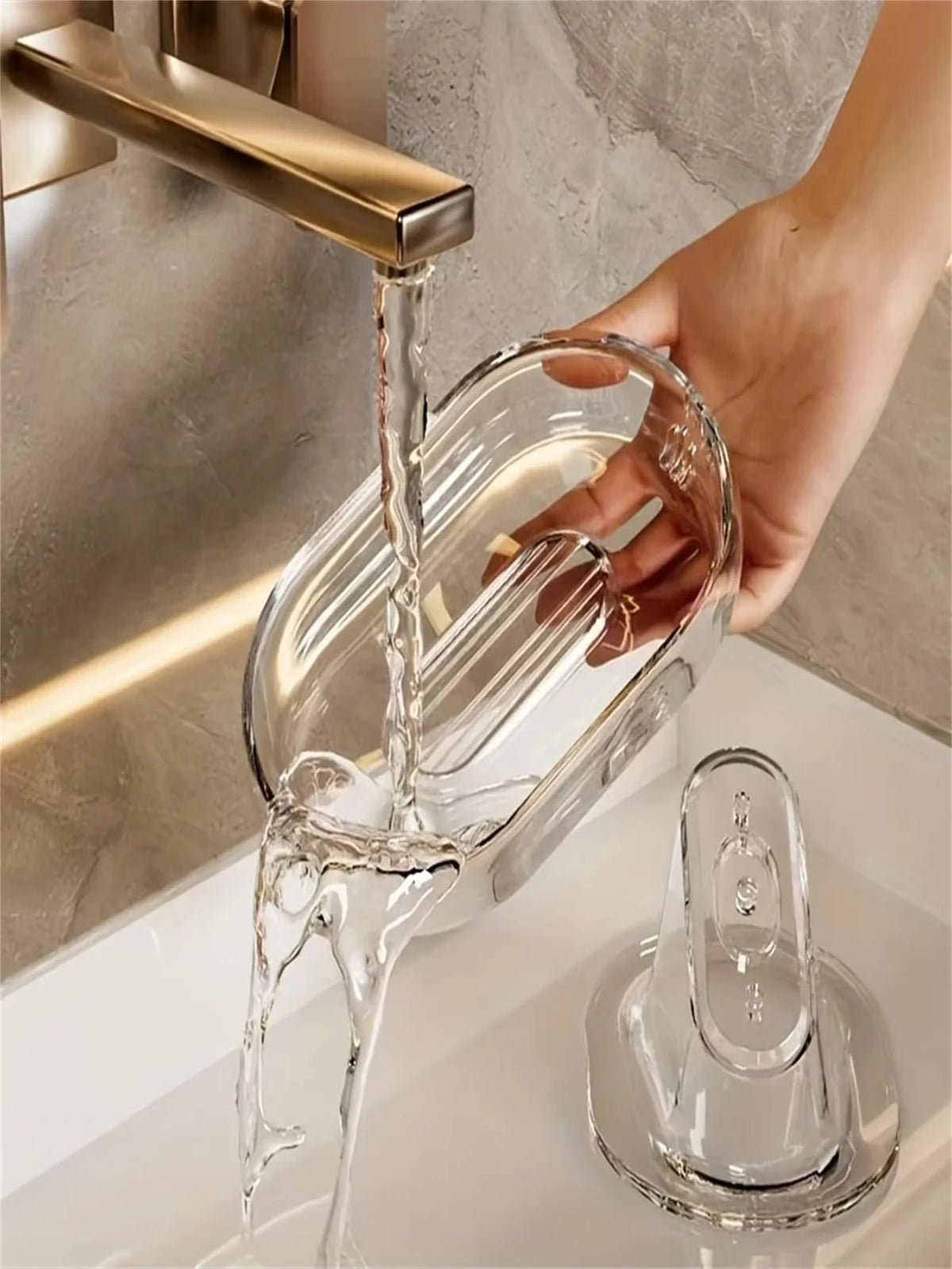 A hand pours clear liquid from a gold faucet into an elegant glass soap dish on a white countertop.