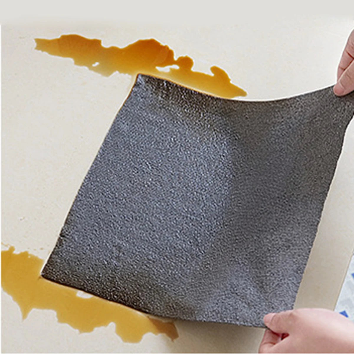 A dark gray reusable multi-surface cleaning wipe is being used to wipe up spilled orange liquid on a light surface.