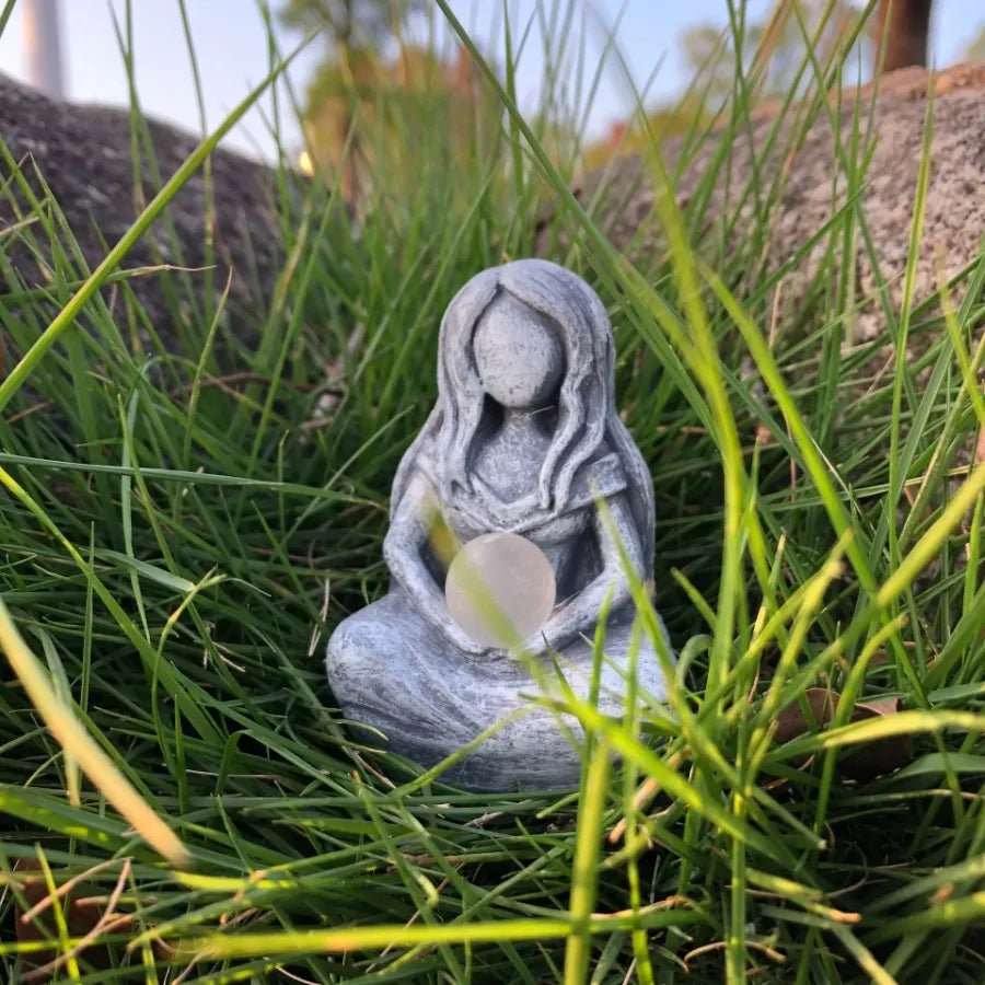 A small gray resin statue of a female figure with long hair sitting in green grass.