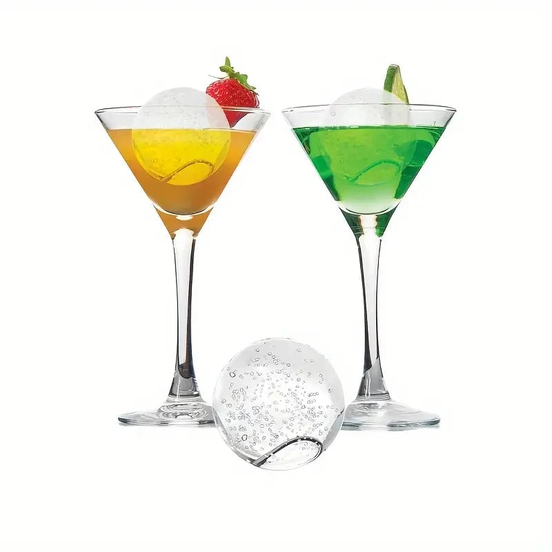 Two martini glasses filled with colorful cocktails garnished with fruit and a floating ice sphere