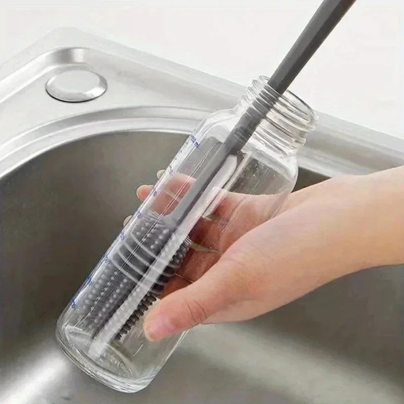 A hand is holding a clear silicone bottle brush with a long handle inside a stainless steel sink for cleaning.