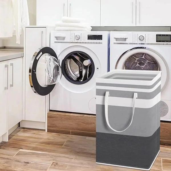 A large foldable canvas laundry hamper with handles sits in front of white washing machines.